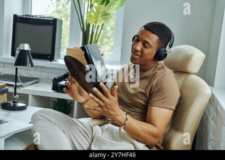 Jeune homme multiracial dans les écouteurs examinant le disque vinyle tout en étant assis sur le lieu de travail Banque D'Images