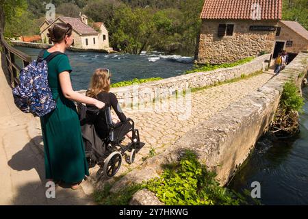 Pousser un enfant handicapé adolescent touriste / fille enfant âgé de 14 ans dans un fauteuil roulant et légèrement en difficulté avec la surface inégale au parc national de Krka, à un point de vue, avec sa mère / maman. Croatie. (138) Banque D'Images