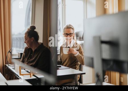 Femme d'affaires mature souriante tenant la tasse tout en étant assis au bureau dans le bureau Banque D'Images