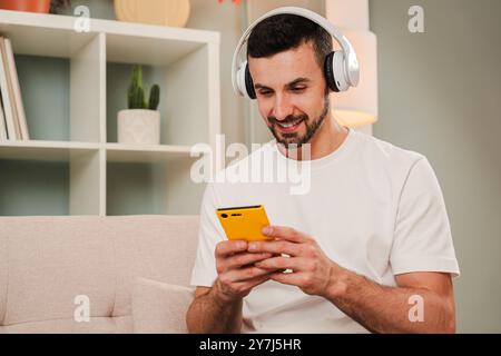 Homme souriant portant des écouteurs à l'aide d'un smartphone jaune à la maison, écoutant probablement de la musique ou des SMS, assis sur un canapé dans une salle détendue, s'engageant avec la technologie et profitant de temps de loisirs. Photo de haute qualité Banque D'Images
