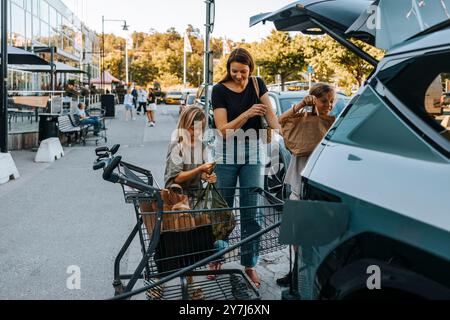 Fils aidant la mère tout en chargeant les sacs d'épicerie dans le coffre de voiture électrique Banque D'Images