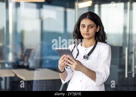 Femme médecin se tient avec tablette et stéthoscope dans le bureau moderne. Professionnel de santé confiant utilisant la technologie pour les consultations médicales, démontrant son expertise dans les solutions de santé numériques. Banque D'Images