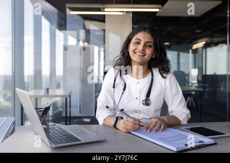 Médecin féminin souriant avec stéthoscope au bureau travaillant avec ordinateur portable, documents et téléphone. Professionnel et confiant praticien médical dans un cadre de bureau moderne se prépare à des tâches importantes. Banque D'Images