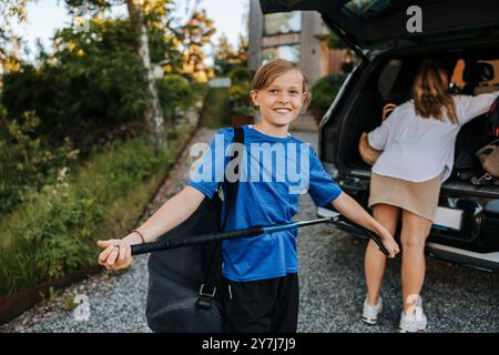 Portrait d'un garçon souriant tenant un bâton de hockey sur glace avec la mère chargeant le coffre de voiture Banque D'Images