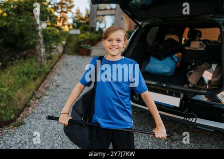 Portrait d'un garçon souriant tenant un bâton de hockey sur glace tout en se tenant devant le coffre de la voiture Banque D'Images