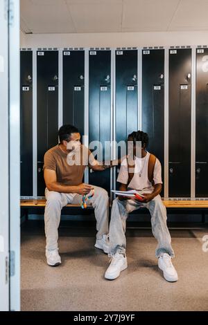 Entraîneur masculin consolant un adolescent assis sur un banc dans un vestiaire à l'école Banque D'Images