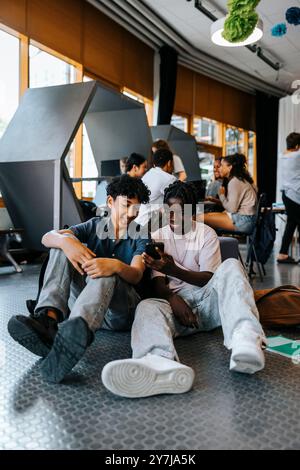 Heureux amis masculins utilisant un téléphone intelligent tout en étant assis dans la cafétéria à l'école Banque D'Images