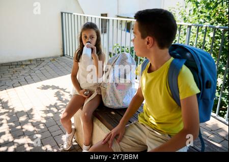 Deux enfants avec des sacs à dos font une pause, assis à l'extérieur et dégustant un verre, sous un ciel ensoleillé. Banque D'Images