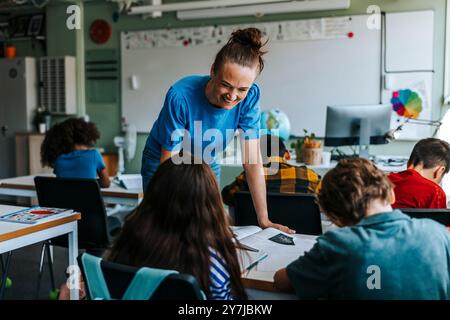 Heureuse professeure pliée tout en parlant avec un élève en classe à l'école primaire Banque D'Images