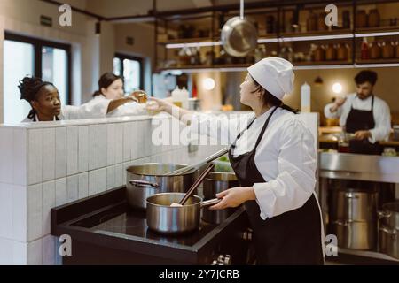 Jeune chef transmettant l'ingrédient au chef féminin préparant la nourriture dans la cuisine commerciale au restaurant Banque D'Images