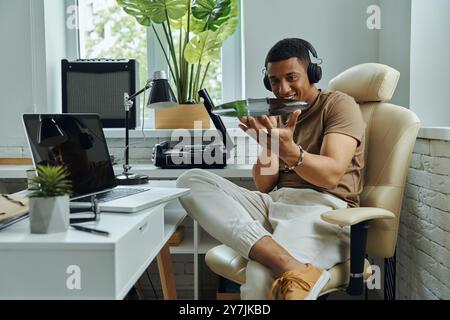 Joyeux homme multiracial dans les écouteurs examinant le disque vinyle tout en étant assis sur le lieu de travail Banque D'Images