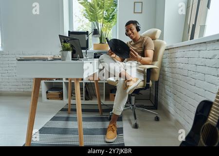 Homme multiracial heureux dans les écouteurs examinant le disque vinyle tout en étant assis sur le lieu de travail Banque D'Images