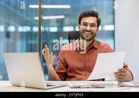 Homme d'affaires confiant souriant tout en tenant le document au bureau. Fait preuve d'énergie positive et de professionnalisme. L'homme s'engage avec la paperasse à côté de l'ordinateur portable, démontrant le lieu de travail Banque D'Images