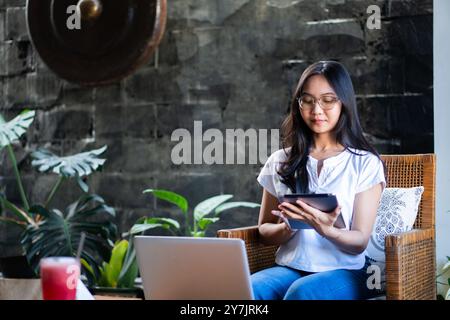 Une jeune femme assise confortablement dans un cadre intérieur confortable, à l'aide d'une tablette. Elle a les cheveux longs et porte des lunettes, habillée d'un haut blanc décontracté et blu Banque D'Images