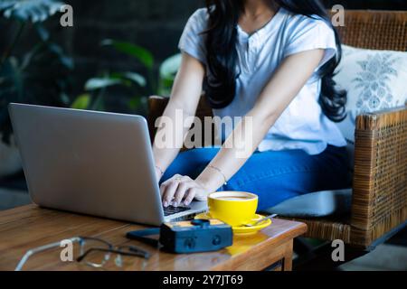 Une femme assise sur une chaise en osier, travaillant sur un ordinateur portable. Elle a les cheveux longs et porte un haut bleu clair décontracté et un Jean. Une tasse de café jaune est posée o Banque D'Images