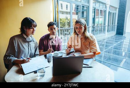 Trois étudiants participent à une session d'étude de groupe sur le campus, en se concentrant sur le travail académique avec des ordinateurs portables et des cahiers dans un environnement lumineux et moderne. Banque D'Images