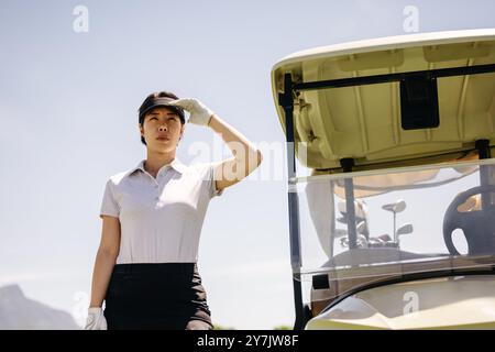 Une femme déterminée golfeuse protège ses yeux du soleil à côté d'une voiturette de golf, participant à une compétition sous un ciel clair. Banque D'Images