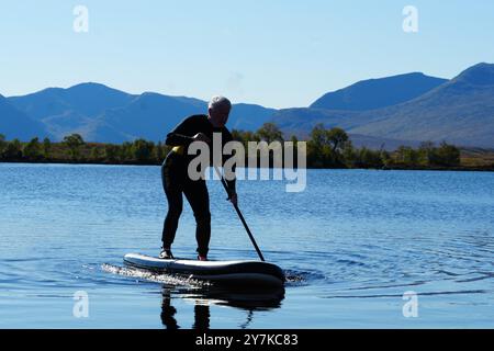 Homme apprenant à paddle board (SUPB) sur le Loch Laidon, avec en toile de fond les montagnes Glen Coe, Rannoch Moor, Écosse, Royaume-Uni Banque D'Images