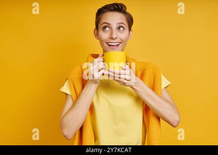 Heureuse jeune femme tenant une tasse avec une boisson chaude tout en se tenant sur fond jaune Banque D'Images