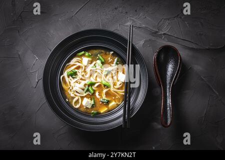 Soupe miso traditionnelle japonaise avec nouilles udon, fromage tofu, poulet, oignons verts et graines de sésame dans un bol noir avec des baguettes et une cuillère sur un Banque D'Images