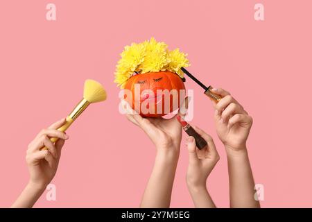 Femmes tenant la citrouille drôle avec le masque à lèvres, les fleurs, les cosmétiques décoratifs et le pinceau de maquillage sur fond rose Banque D'Images