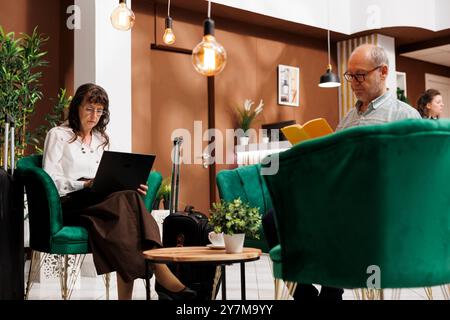 Vieille femme retraitée avec des écouteurs, assise sur un canapé dans le hall de l'hôtel et utilisant un ordinateur portable. Un homme lisant un livre alors qu'une dame âgée avec un micro-casque sans fil utilise son mini-ordinateur dans le salon. Banque D'Images