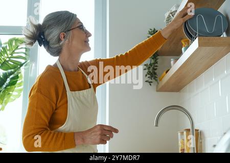 Femme aînée confiante mettant des assiettes sur l'étagère tout en se tenant debout à la cuisine domestique Banque D'Images