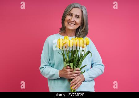 Femme âgée heureuse tenant un bouquet de tulipes tout en se tenant debout sur fond rose Banque D'Images