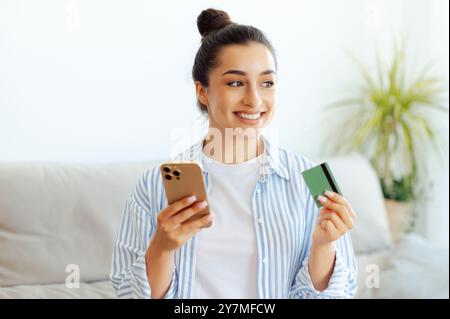 Concept e-banking. Joyeuse jolie femme brune, assise sur un canapé dans un salon, tient smartphone et carte de crédit pour le paiement en ligne dans une application mobile, faire des achats en ligne, regarder loin, sourire Banque D'Images