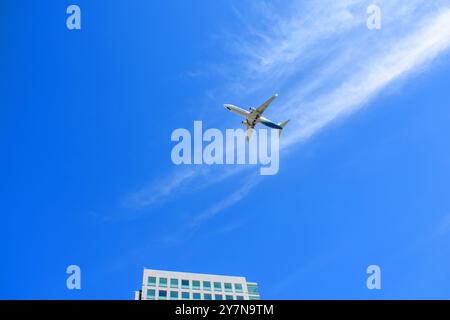 Avion Alaska Airlines volant contre un ciel bleu vibrant avec des nuages tortueux, passant au-dessus d'un immeuble de bureaux moderne - San Jose, Californie, États-Unis - sept Banque D'Images