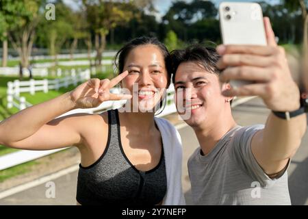 Un jeune couple asiatique heureux en vêtements de sport prend un selfie ensemble après avoir fait du jogging dans un parc par une journée ensoleillée. activité de plein air, sport, modes de vie Banque D'Images