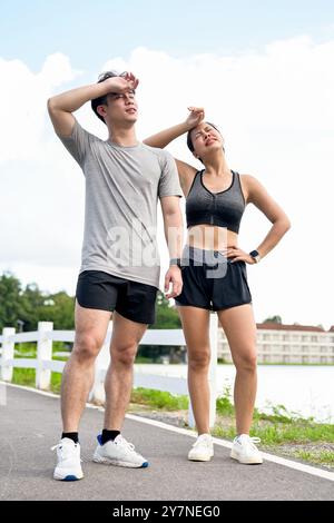 Un couple asiatique fatigué et en sueur en vêtements de sport essuie la sueur de leurs visages, prenant une pause après une longue course ensemble dans un parc le matin. c.a. extérieur Banque D'Images