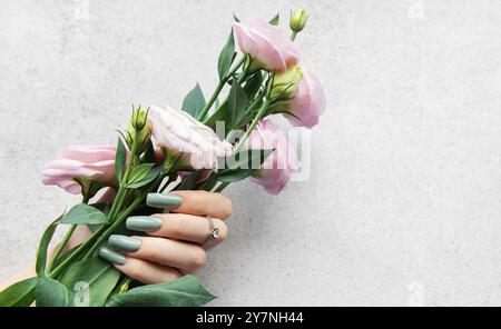 Une main ornée de longs ongles verts doux tient doucement un bouquet de fleurs roses. La toile de fond grise simple souligne la beauté des fleurs et Banque D'Images