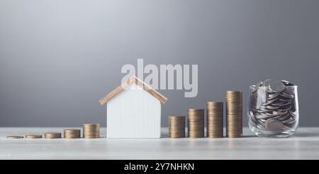 L'homme laisse tomber des pièces de monnaie dans un bocal en verre, avec des piles de pièces de monnaie et un modèle de maison en bois à côté de lui. Souligne le concept d'économiser de l'argent pour la future maison dans Banque D'Images