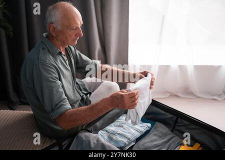 Concept de voyage. Homme de retraite préparant le sac à bagages d'emballage pour le voyage de vacances d'été assis sur le lit à la maison, emballer des vêtements dans la valise pour un nouveau Banque D'Images