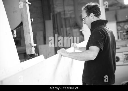 L'image en noir et blanc d'Un charpentier travaille sur un grand panneau de bois dans son atelier, en utilisant une variété d'outils et d'équipements Banque D'Images