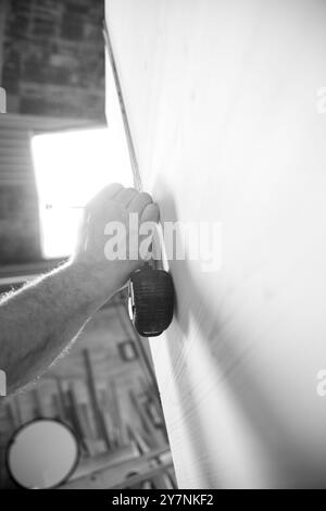 L'image en noir et blanc d'Un charpentier utilise un mètre ruban pour vérifier les dimensions d'une grande planche de bois tout en travaillant dans son atelier Banque D'Images