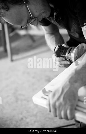 Image en noir et blanc d'Un charpentier ponçant un morceau de bois dans son atelier à l'aide d'une ponceuse électrique Banque D'Images