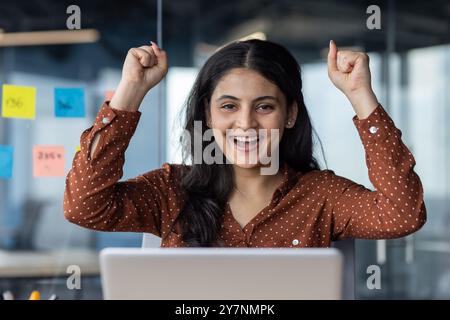 Joyeuse jeune femme lève les bras pour célébrer le succès dans l'environnement de bureau. L'expression enthousiaste transmet le bonheur et la réussite. Le fond présente un mur de verre avec des notes adhésives. Banque D'Images