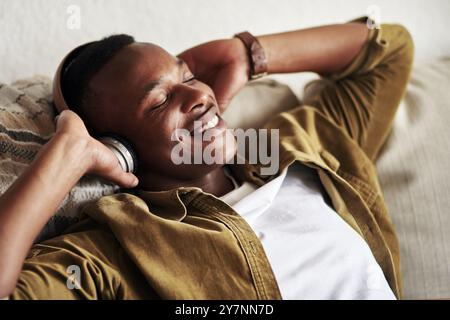 Heureux, homme noir et détendez-vous avec des écouteurs sur le canapé pour la musique, l'écoute ou podcast à la maison. Jeune Africain, personne masculine ou utilisateur avec sourire ou casque Banque D'Images