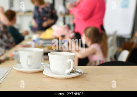 Deux tasses à thé blanches sur une table en bois avec un fond flou de personnes en réunion. Table en bois clair, surface lisse, tasses design simples, petite cuillère. Banque D'Images