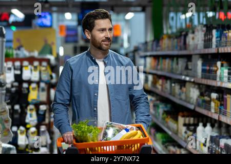 Homme magasinant dans un supermarché poussant chariot orange rempli de produits d'épicerie. L'accent est mis sur les produits frais comme la laitue et les bananes. Scene capture l'expérience quotidienne de magasinage et le style de vie des consommateurs. Banque D'Images