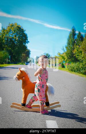 Une petite fille est seule à côté d'un cheval jouet en plein air au milieu de la route Banque D'Images