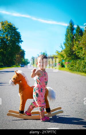 Une petite fille est seule à côté d'un cheval jouet en plein air au milieu de la route Banque D'Images