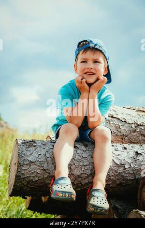 Un petit garçon portant une casquette de baseball bleue et un short est assis sur une bûche et regarde au loin Banque D'Images