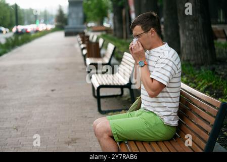 Allergies saisonnières ou rhume avec un nez qui coule. Un homme portant une chemise rayée et un short vert est assis sur un banc dans un parc, éternuant dans un mouchoir par une journée calme. Banque D'Images