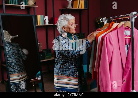 Femme âgée de cheveux gris à la mode examinant des vêtements suspendus sur des supports dans la boutique de mode Banque D'Images