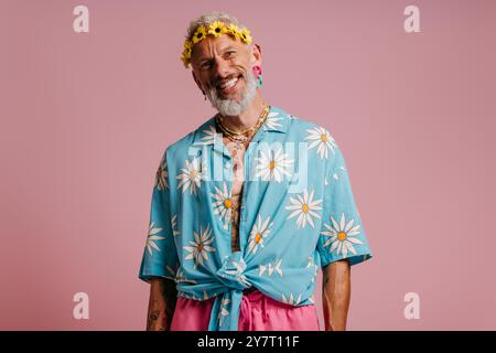 Heureux homme senior dans la chemise élégante et la couronne de fleurs sur la tête debout sur fond rose Banque D'Images