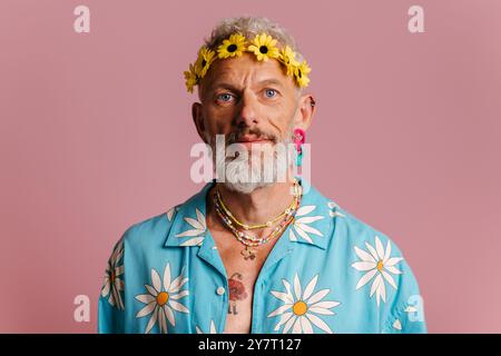 Bel homme senior en chemise élégante et couronne de fleurs sur la tête debout sur fond rose Banque D'Images