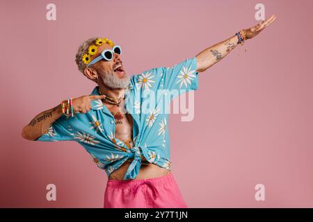 Homme senior à la mode en couronne florale pointant loin et souriant sur fond rose Banque D'Images